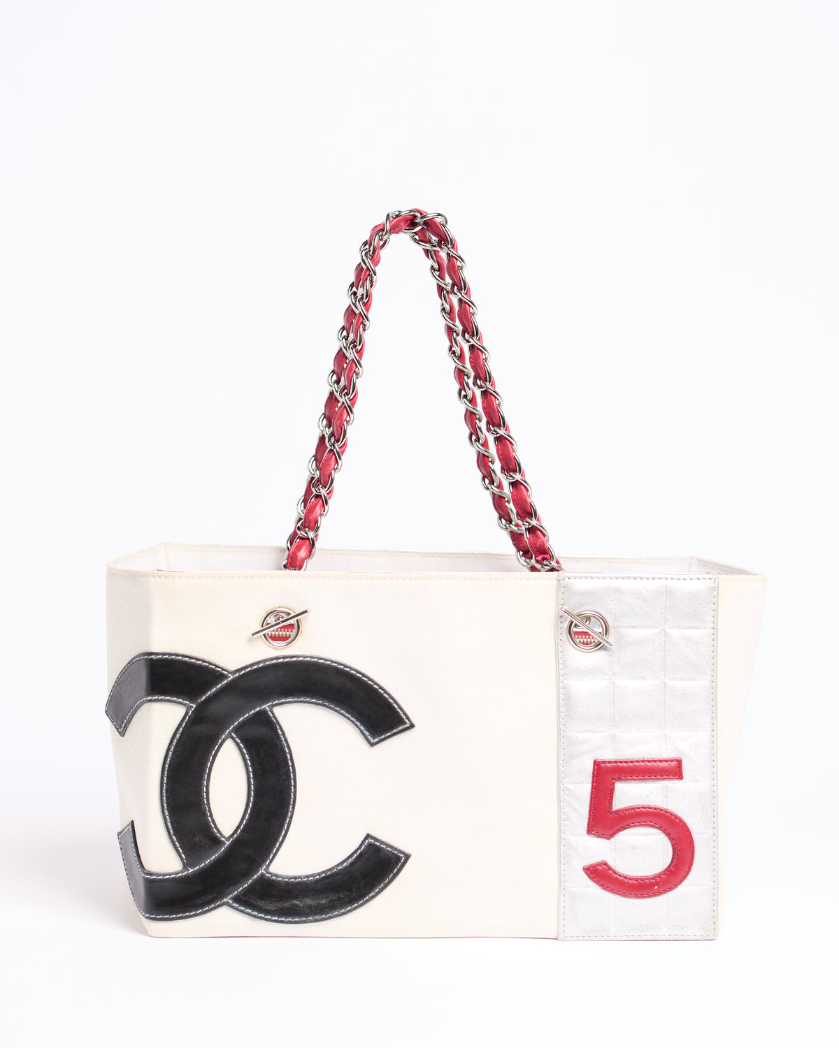 A Chanel tote bag in off-white canvas with a black interlocking CC logo, a red textured '5' on the side, and red-and-silver chain handles, shown against a plain white background.